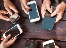 Photo of a set of hands belonging to multiple individuals holding cell phones on a picnic table.